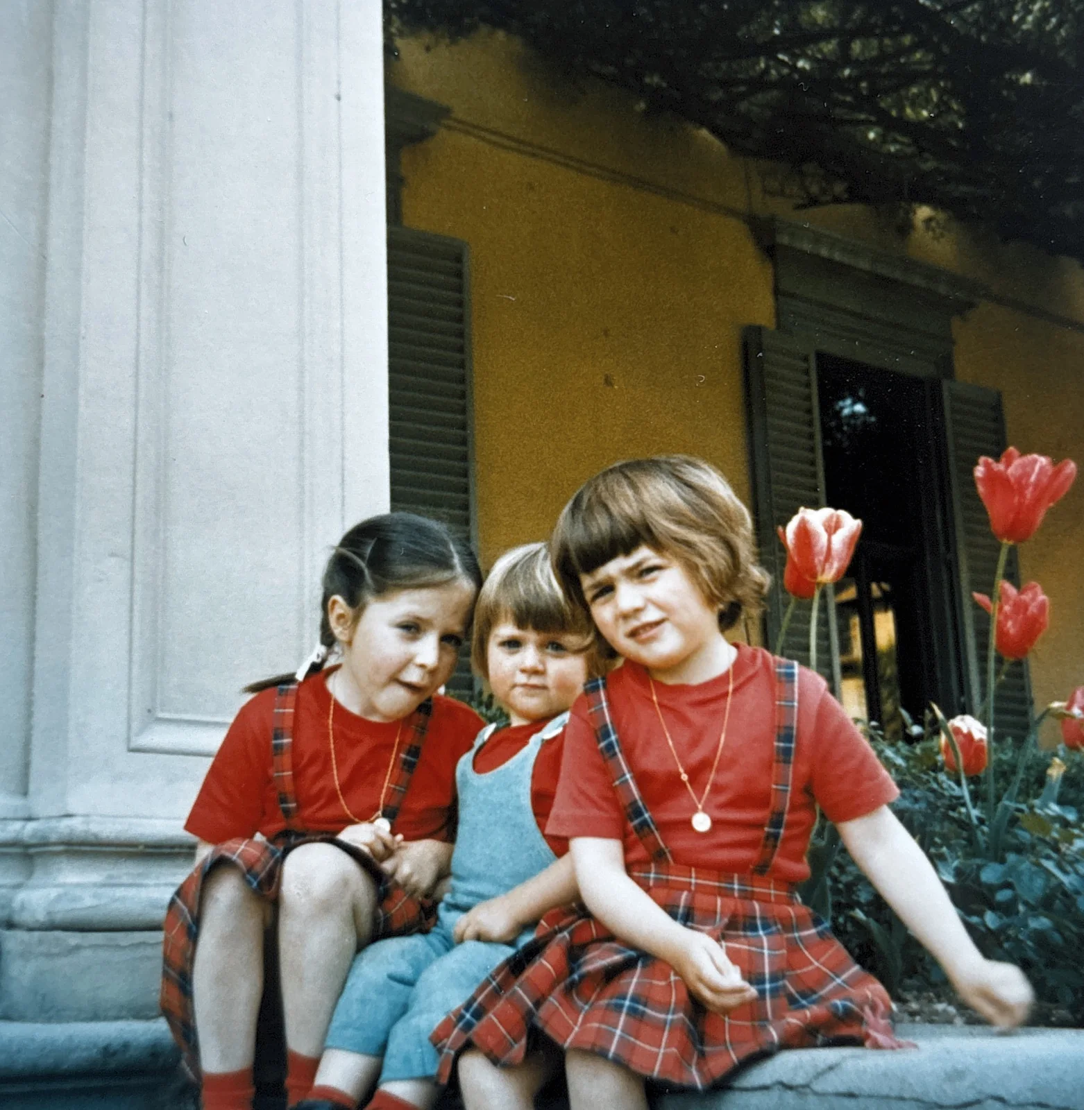 Photographie d’archive : trois enfants assis sur les marches d’une maison, près de tulipes rouges