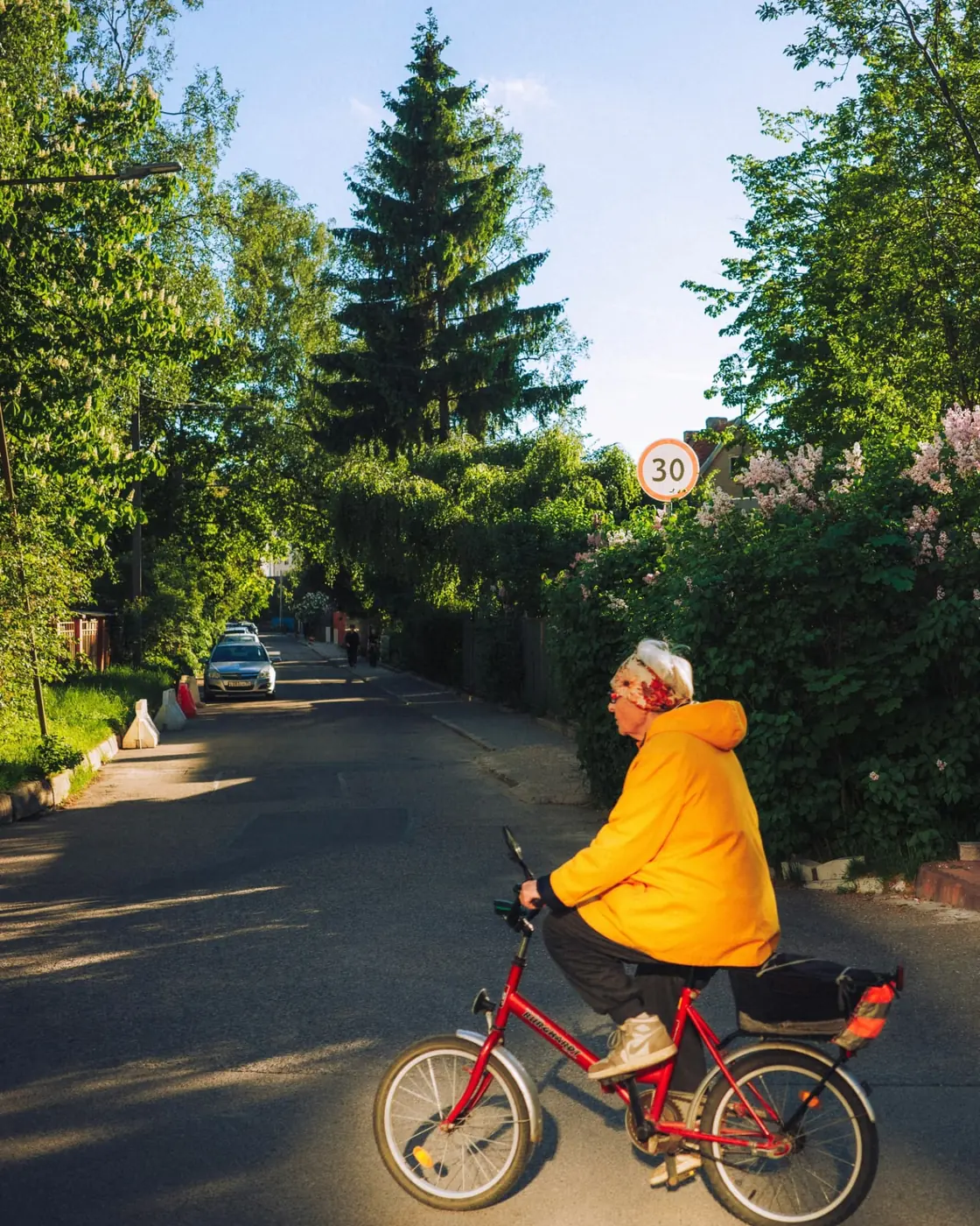 Une femme âgée en veste jaune pédale sur un vélo rouge le long d'une rue ensoleillée bordée de verdure