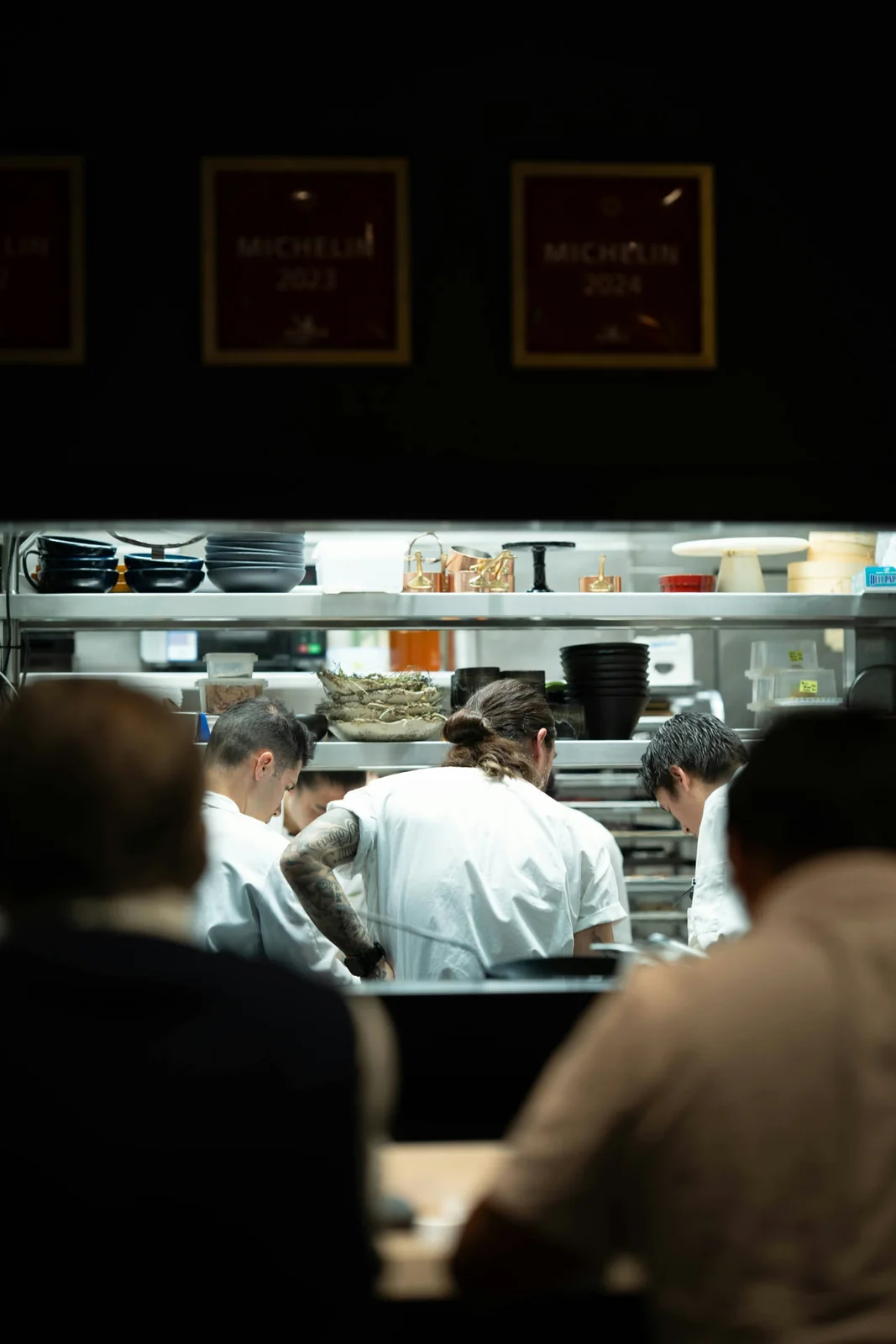 Vue plongeante sur une brigade de cuisine étoilée au travail, à contre-jour, encadrée par les silhouettes de convives au premier plan